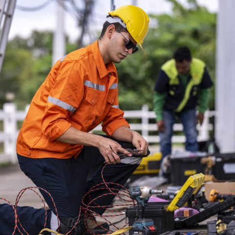 A skilled technician in a bright orange safety jacket and hard hat prepares electrical wiring with precision. Tools and open cases are spread in the background, showcasing a professional workspace.
