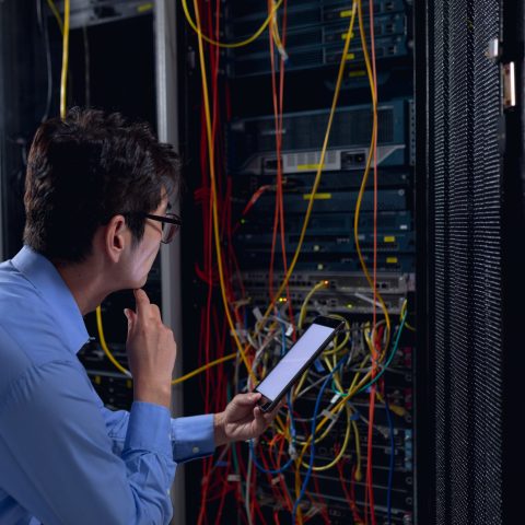 Thoughtful asian male engineer using digital tablet and inspecting computer server in server room. database server management and maintenance concept